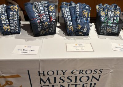 Display table with memorabilia, booklets, stickers, and tshirts with the Holy Cross Mission Center logo.