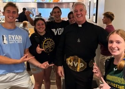 Fr. Tom Eckert, C.S.C., wearing an MVP wrestling belt, surrounded by University of Notre Dame students.